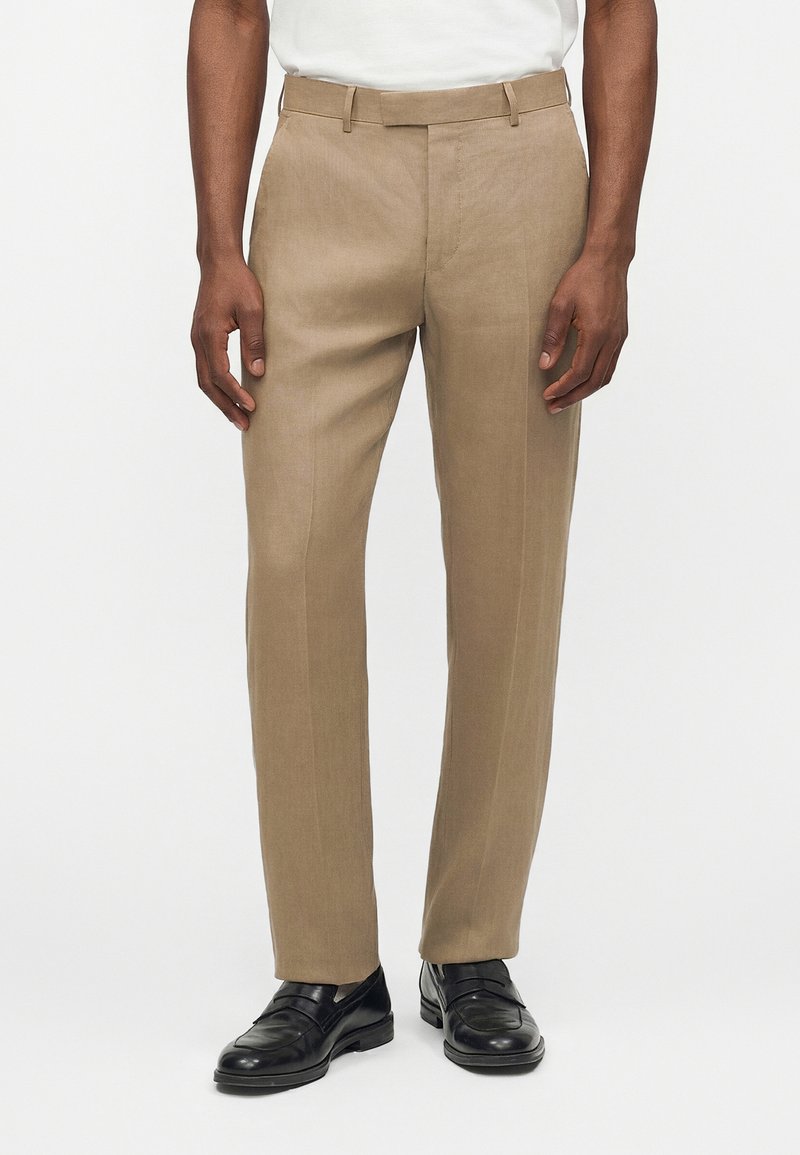 Μπεζ ραμμένο παντελόνι με πτυχή μπροστά, θηλιές για ζώνη, φορεμένο με μαύρα δερμάτινα μοκασίνια και λευκό πουκάμισο μέσα στην παντελόνα.