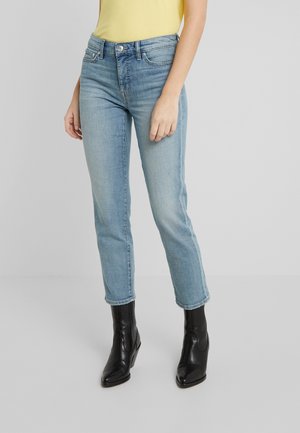 Femme portant un jean droit bleu clair, un haut jaune et des bottines noires à talons, debout devant un fond uni.