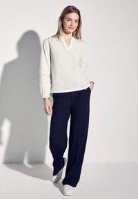 Vit långärmad blus med V-ringning och samlade handledsdetaljer, kombinerad med lösa marinblå byxor och vita sneakers. En enkel, modern design.