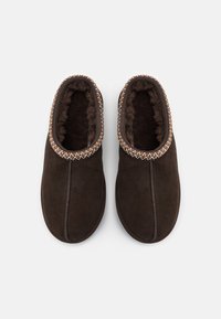 Chaussons en suede marron avec une doublure intérieure en peluche, ornés d'une finition tressée décorative autour de l'ouverture et d'un design lisse et arrondi.