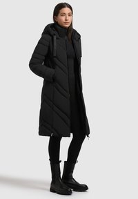 En ung kvinde står iført en lang sort quilted frakke med hætte, sorte leggings og sorte læderstøvler foran en ensfarvet lys baggrund.