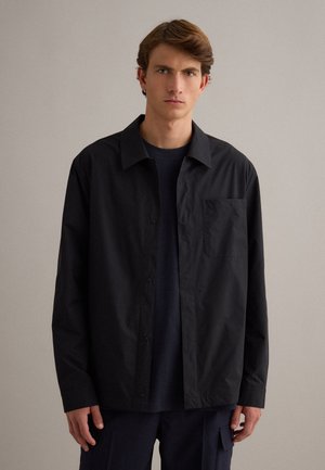 Giovane uomo con capelli castano chiaro che indossa una camicia nera con bottoni sopra una maglietta scura, in piedi davanti a uno sfondo chiaro e semplice.