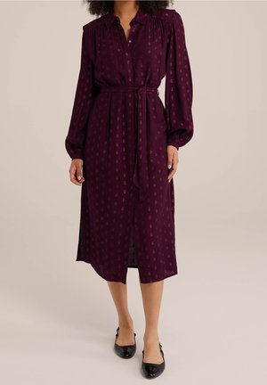 Femme portant une robe midi à manches longues, boutonnée, de couleur violet foncé avec une ceinture à la taille et un petit motif imprimé, associée à des chaussures noires.