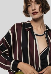 Blusa di seta con righe verticali color bordeaux e blu navale, design con colletto e scollatura a V profonda. Presenta una collana argentata e maniche arrotolate.