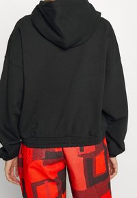 Felpa nera con cappuccio regolabile, maniche ampie e orlo inferiore elasticizzato. Indossata con pantaloni a fantasia rossi brillanti. Tessuto morbido e casual.