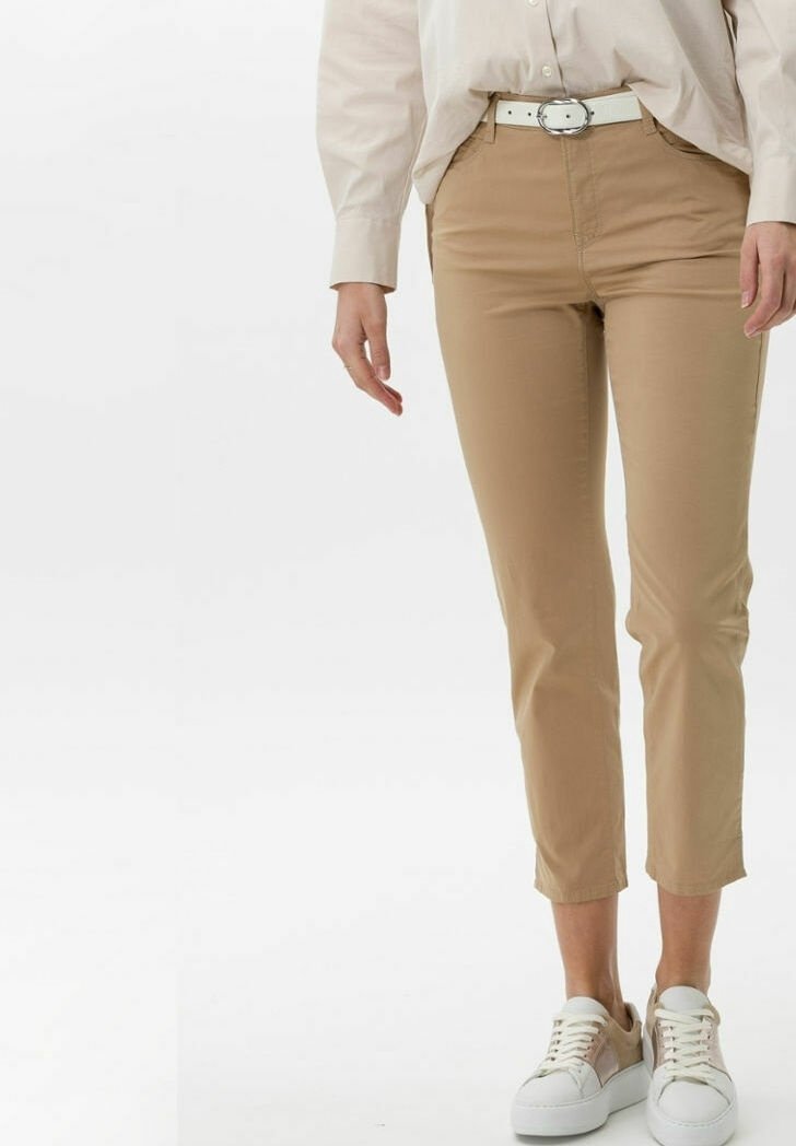 Femme portant un pantalon beige court, une ceinture blanche, un chemisier crème et des baskets blanches, debout devant un fond uni.