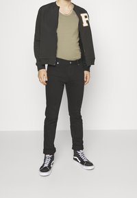 Chaqueta bomber negra con cremallera y una gran letra "R" de fieltro, camiseta sin mangas oliva, jeans ajustados negros y zapatillas altas negras con suelas blancas.