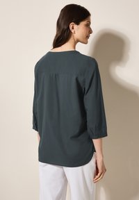 Dunkelbluse in Teal mit Dreiviertelärmeln, rundem Ausschnitt und gefaltetem Rückendetail. Kombiniert mit weißen Hosen, die eine glatte Textur aufweisen.