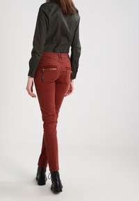 Red slim-fit pants with zipper pockets and black accents, paired with a dark green collared shirt. The outfit features a fitted design.