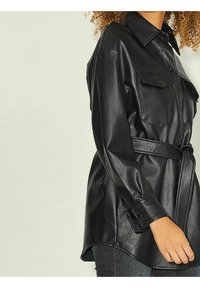 Mustat nahka-paitajakku vyöllä, jossa on kaksi rintataskua, nappikiinnitys ja pitkät hihat. Se on valmistettu sileästä materiaalista ja siinä on klassinen kaulus.