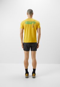 Gelbes Baumwoll-T-Shirt mit türkisfarbenem Text und einem Grafikdesign auf dem Rücken, kombiniert mit schwarzen Shorts und mehrfarbigen Sneakers.