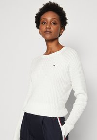 Maglione bianco lavorato a maglia con un motivo testurizzato, orlo a coste e scollatura rotonda. Presenta un piccolo logo sul petto.