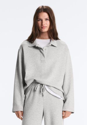 Młoda kobieta o długich brązowych włosach, ubrana w jasnoszarą koszulkę polo z długim rękawem oraz pasujące spodnie z troczkiem, na białym topie.