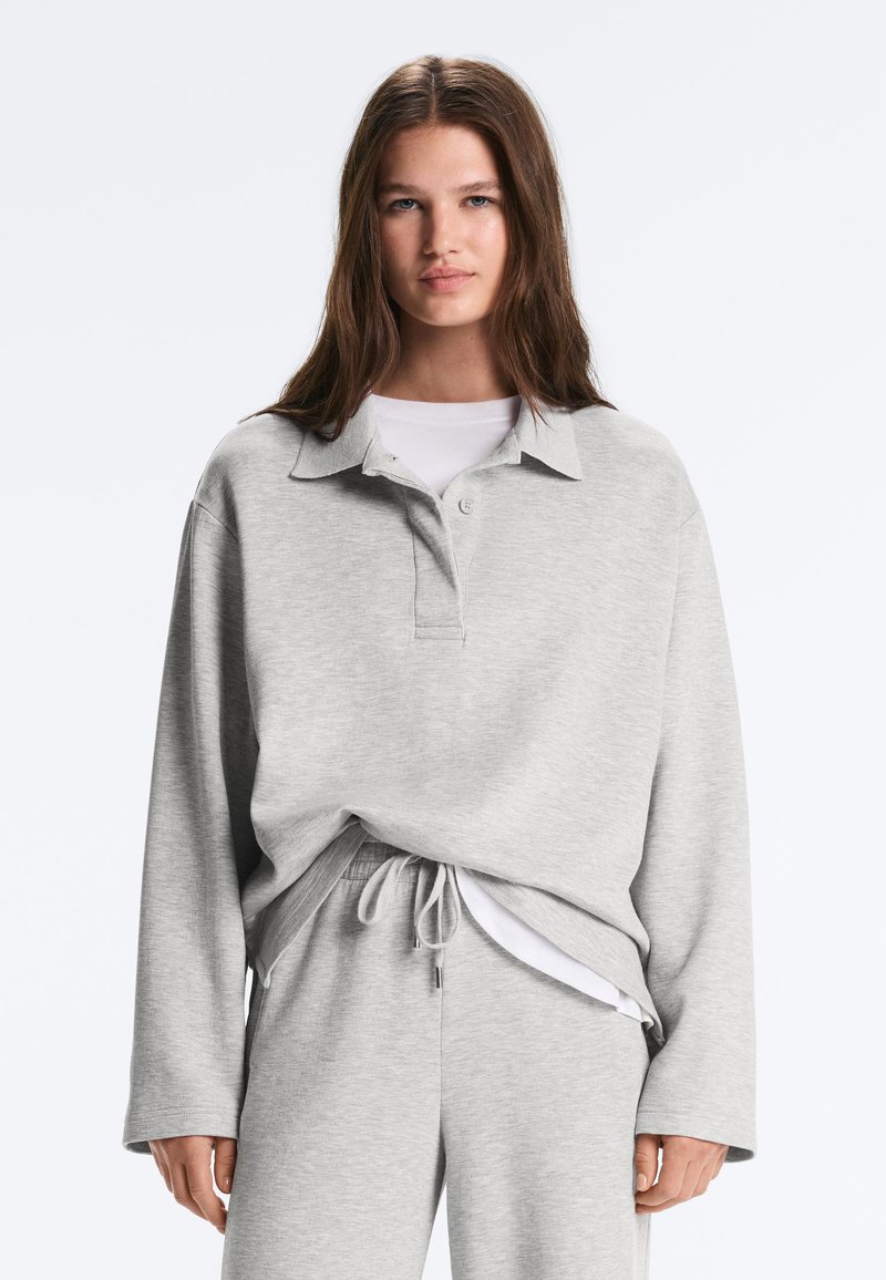 Jeune femme aux longs cheveux bruns portant un polo à manches longues gris clair et un pantalon à cordon assorti sur un haut blanc.