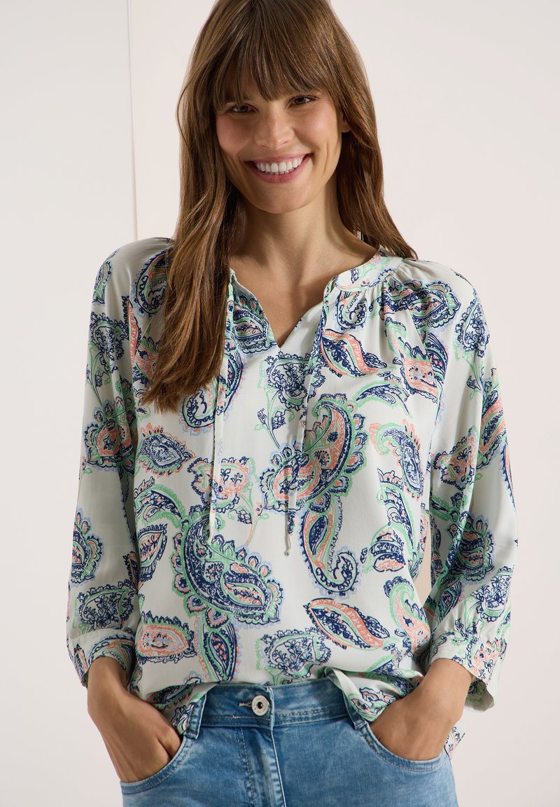 Femme souriante aux longs cheveux bruns portant un chemisier blanc à motif cachemire et un jean bleu, les mains dans les poches, debout devant un fond uni.
