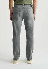 Jeans de mezclilla gris con corte recto, que cuentan con bolsillos traseros y un parche de cuero negro en la cintura. Textura suave y detalles mínimos.