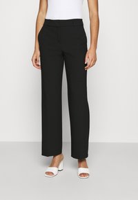 Pantalones negros de pierna ancha con un diseño entallado, que presentan una textura suave y una cintura plana. Combinados con sandalias blancas de tacón.