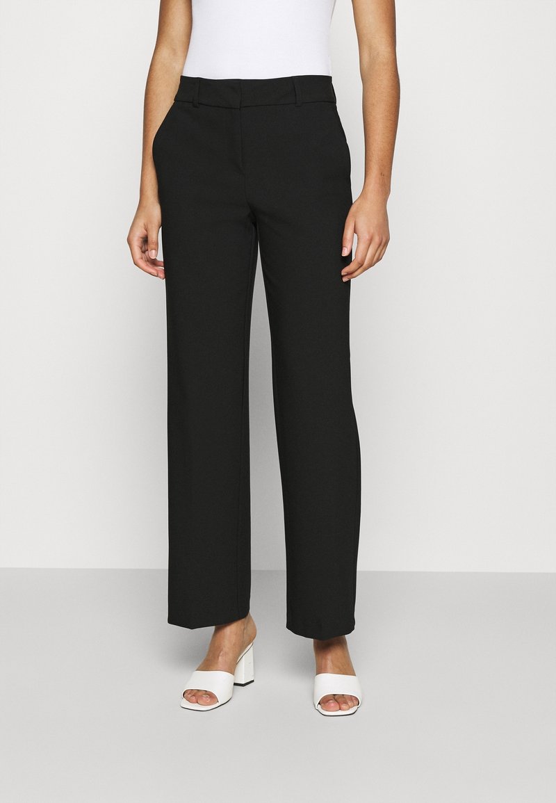 Pantalones negros de pierna ancha con un diseño entallado, que presentan una textura suave y una cintura plana. Combinados con sandalias blancas de tacón.