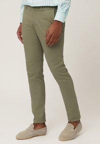 Olive green chinos made of cotton with a slim fit, flat front, and side pockets, paired with gray slip-on shoes.