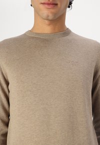 Μπεζ ελαφρύ πουλόβερ με στρογγυλή λαιμόκοψη, ριμπ μανσέτες και μια διακριτική λογότυπη στο στήθος. Λείος υφασμάτινος χαρακτήρας, εφαρμοστή σχεδίαση.