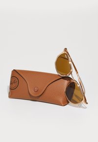 Ochelari de soare Ray-Ban cu lentile maro, ramă din metal auriu și carcasă din piele texturată maro, care prezintă un logo și închidere cu buton.