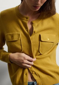 Cardigan giallo senape con scollatura rotonda, due tasche anteriori e chiusura con bottoni; realizzato in un tessuto a maglia texturizzato.