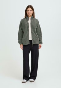 Grøn, fluffy fleecejakke med høj krave, båret over en lys sweater, parret med sorte bukser med vidde og grå slip-on sko.