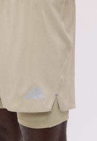 Shorts de sport beige avec leggings de compression intégrés et une petite fente latérale, avec un logo réfléchissant près de l’ourlet.