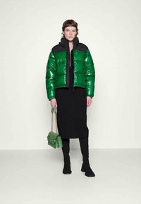 Casaco puffer verde com um acabamento brilhante, detalhes em preto e fecho zip. Combinado com uma saia preta, botas até ao joelho e uma mala verde.