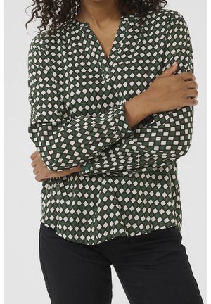 Femme portant un chemisier à manches longues à motif losanges verts et blancs avec poignets boutonnés, associé à un pantalon noir, bras croisés.