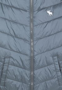 Giacca imbottita grigia con zip frontale, due tasche laterali e un piccolo emblema di alce bianco sul lato sinistro del petto.