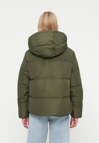 Persoană blondă îmbrăcată cu o geacă cu glugă de culoare verde măslinie și blugi de culoare albastră deschisă, shownă de la spate pe un fundal alb.