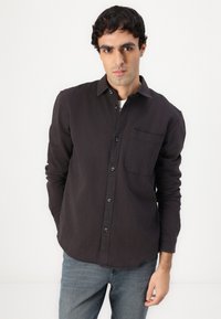 Camicia grigia scura con bottoni, realizzata in tessuto leggero, con una tasca sul petto e bottoni neri. Indossata con jeans blu per creare contrasto.