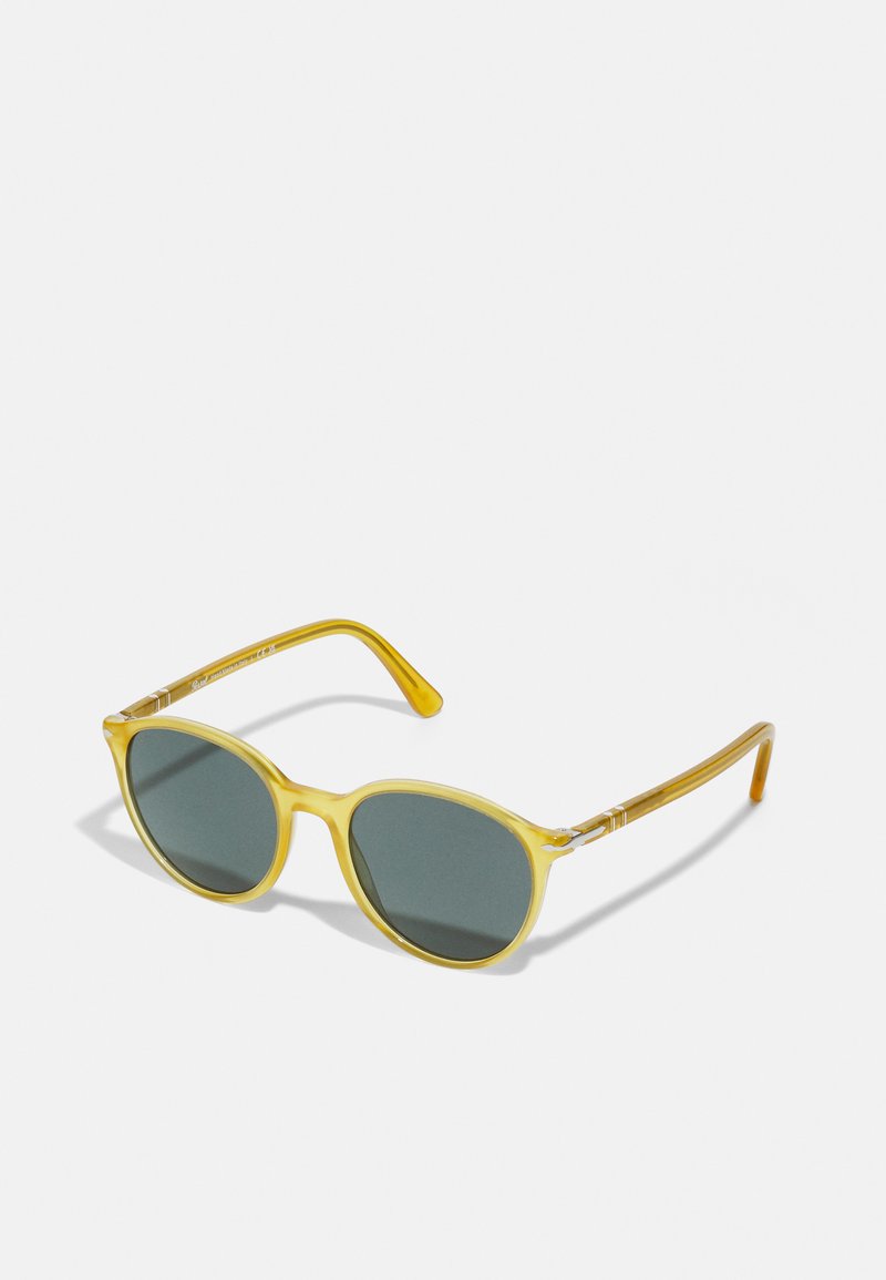 Lunettes de soleil jaunes transparentes avec des verres verts, monture arrondie et branches fines comportant des accents métalliques subtils.
