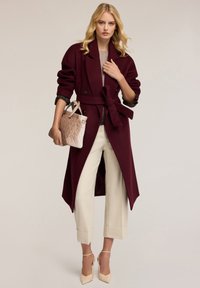Cappotto trench in lana bordeaux con cintura in vita, abbinato a pantaloni cropped crema e tacchi beige, mentre tiene una borsa in pelle di un rosa chiaro testurizzato.