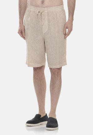 Uomo che indossa pantaloncini beige con coulisse al ginocchio e scarpe slip-on nere, in piedi davanti a uno sfondo chiaro e semplice.