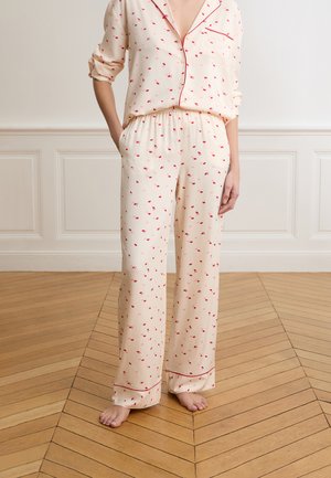 Kobieta w kremowej piżamie z czerwonymi i różowymi nadrukami ust, stojąca boso na drewnianej podłodze, z jedną ręką w kieszeni.