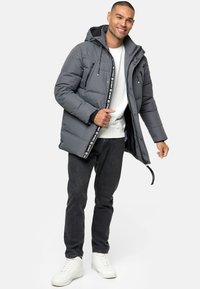 Grå pufferjakke med hætte, med lynlåslommer på brystet, logo-detaljer og lynlås ved fronten. Bæres over en hvid skjorte og sorte jeans.