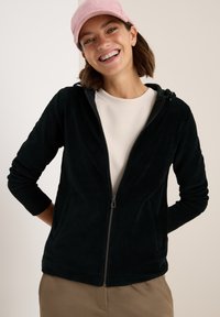 Haut zippé en velours côtelé noir avec une capuche et deux poches avant. Porté sur une chemise claire, associé à un pantalon kaki. Casquette rose sur la tête.