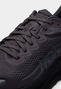 Κοντινή λήψη σκούρου γκρι αθλητικού παπουτσιού με υφή διχτυού, χοντρές κορδόνια και λογότυπο "HOKA" στο πλάι.