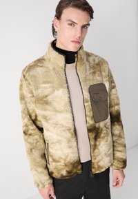 Fleecejack in een tan en groen camouflagedesign, met een ritssluiting aan de voorkant, een staande kraag en een voorzak met een drukknoopsluiting.