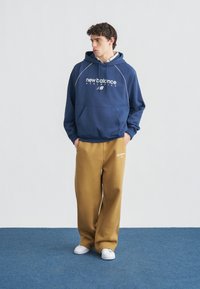 Jonge man met een marineblauwe New Balance-trui en beige joggingbroek, staand met handen in de zakken op een blauw tapijt.