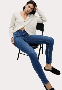 Vit kortärmad kavaj med knäppning och fickdetaljer, i kombination med blå tajta jeans. Modellen sitter på en svart stol, med svarta skor.