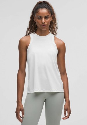 A woman with curly hair models a sleeveless white top and light green leggings, standing in front of a gray background.