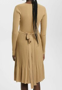 Långärmad plisserad klänning i beige ribbad stickning, med en figurnära bodice och en knytning vid midjan bak, samt en utsvängd nederdel.