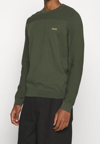 Dunkelgrüner Strickpullover mit gerippten Akzenten auf den Schultern, Rundhalsausschnitt und kleinem goldfarbenem "BOSS"-Logo auf der Brust; figurnaher Schnitt.