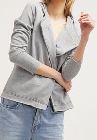 Veste à capuche grise avec des manches longues, devant drapé et coupe décontractée. Fabriquée en tissu doux avec des coutures visibles et un ourlet lâche.