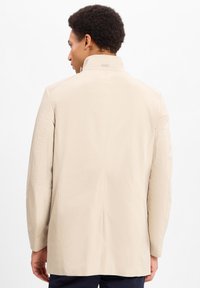 Beige lichte jas met een hoge kraag, rechte snit en minimale stiksels, met een klein logo op de achterkant.