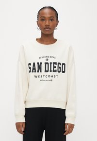Frau trägt ein cremefarbenes Sweatshirt mit dem Aufdruck "SAN DIEGO WESTCOAST", schwarze Hose und Creolen, steht vor weißem Hintergrund.