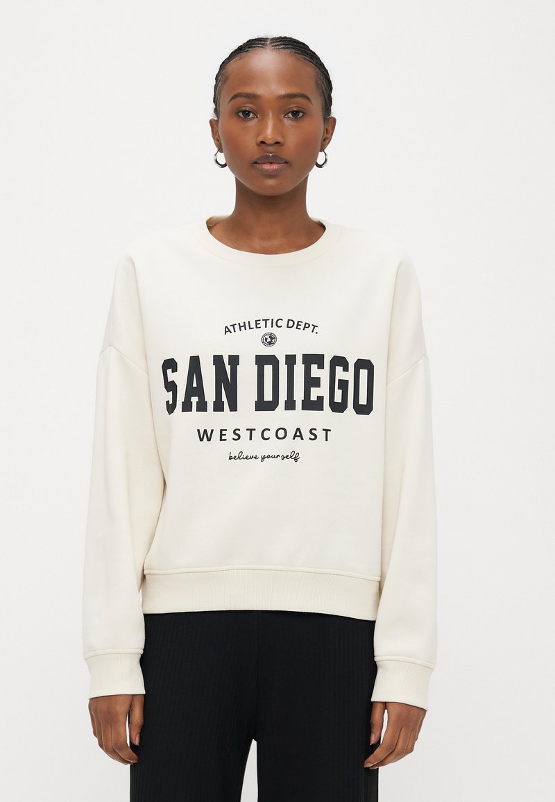 Frau trägt ein cremefarbenes Sweatshirt mit dem Aufdruck "SAN DIEGO WESTCOAST", schwarze Hose und Creolen, steht vor weißem Hintergrund.